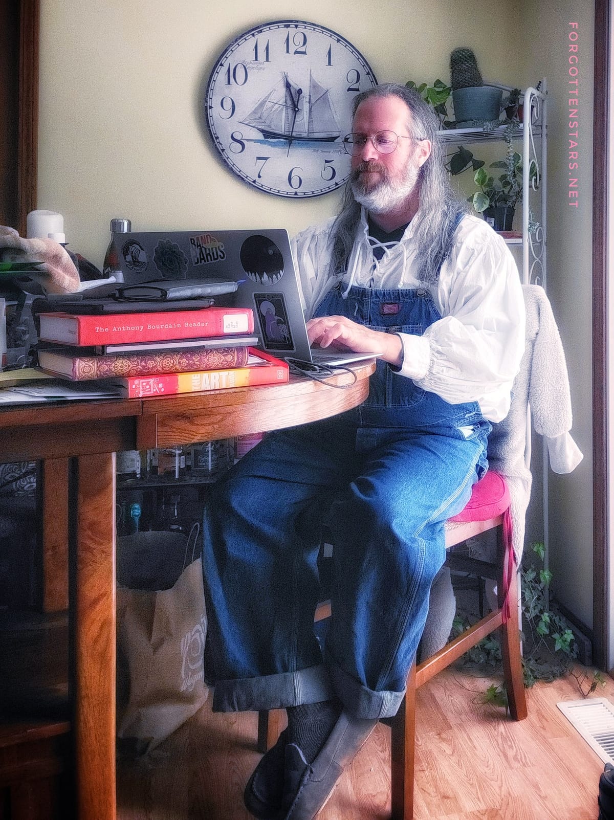 Your longhaired bearded Correspondent, writing at a tall table. Outfit: a white ren-fest style shirt with long and very poofy sleeves, and a pair of vintage blue denim Dickies bib overalls.