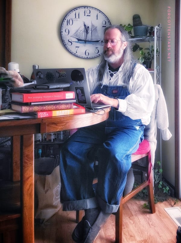Your longhaired bearded Correspondent, writing at a tall table. Outfit: a white ren-fest style shirt with long and very poofy sleeves, and a pair of vintage blue denim Dickies bib overalls.